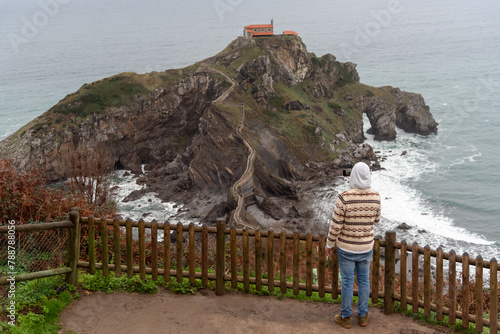 Man with his back turned watching the hermitage of San Juan de Gaztelugatxe located on a hill in the middle of the sea on the touristy coast of Biscay. photo
