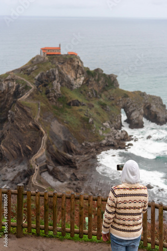 Man with his back turned taking a photo with his mobile phone to the hermitage of San Juan de Gaztelugatxe on a hill in the middle of the sea on the touristy coast of Biscay. photo