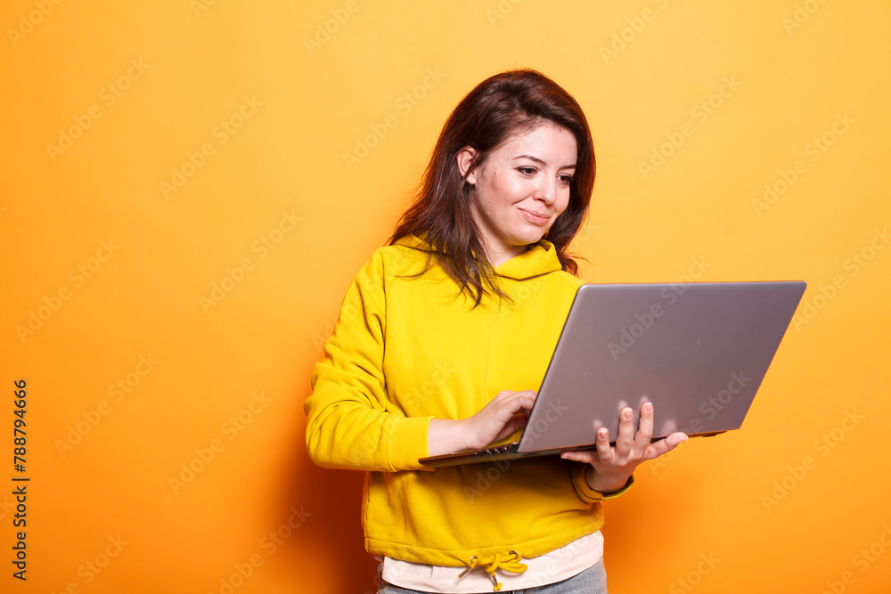 Naklejka premium In the studio, young adult looks at a laptop to use technology on camera. Portrait of a modern woman standing over an isolated background, holding a digital device and surfing the internet.