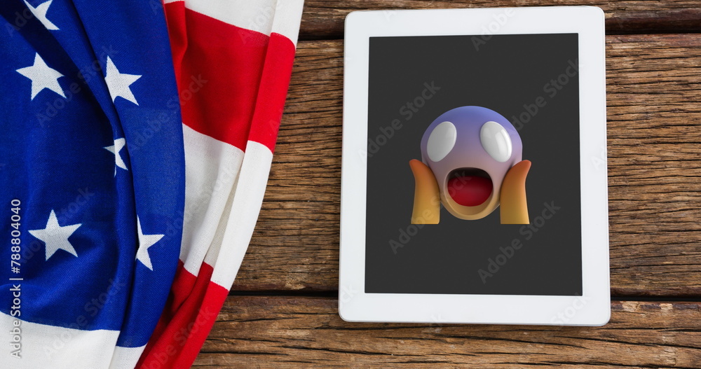 Fototapeta premium A tablet displaying shocked emoji rests on a wooden surface beside American flag