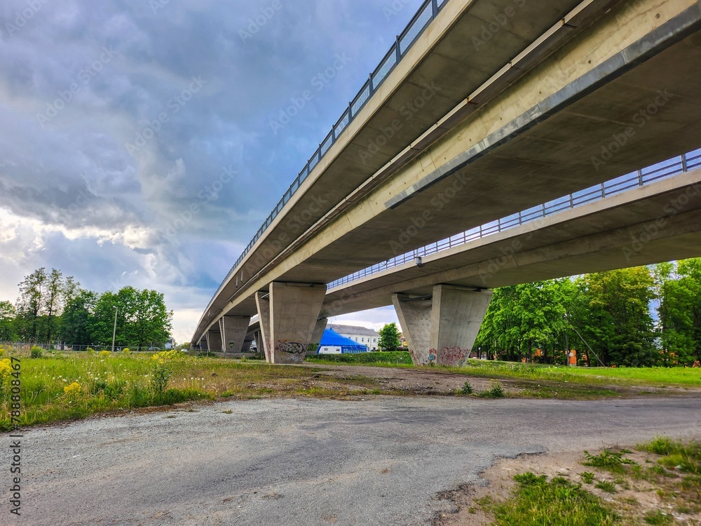 A wide concrete bridge arches gracefully over a bustling road, framed by dense foliage. The overcast sky casts a moody atmosphere, enhancing the industrial urban scene.