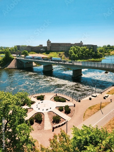 Enchanting view of the Narva City on a sunny day, with a bridge over calm waters reflecting the blue sky, surrounded by lush greenery and vibrant flowers.