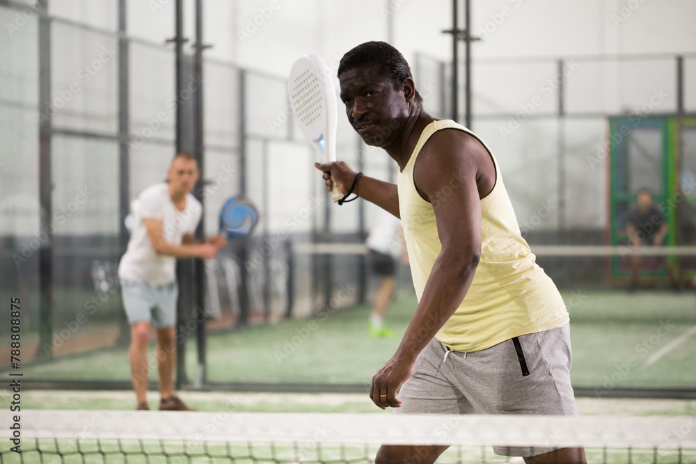 Portrait of sporty adult african american man playing padel on indoor court, ready to hit ball. Sport and active lifestyle concept.