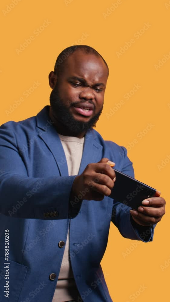 Vertical video BIPOC person playing videogames on smartphone, having negative burst of emotions after defeat. Frustrated gamer losing game on phone, isolated over studio background, camera B Vertical video BIPOC person playing videogames on smartphone, having negative burst of emotions after defeat. Frustrated gamer losing game on phone, isolated over studio background, camera B