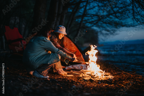 Two friends roast marshmallows over a cozy bonfire by the lake at dusk, showcasing a serene camping experience.