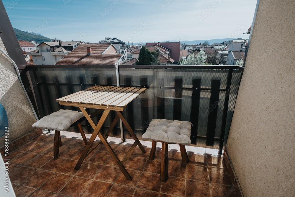Obraz premium A comfortable small balcony featuring a wooden table and cushioned chairs with a scenic urban backdrop.