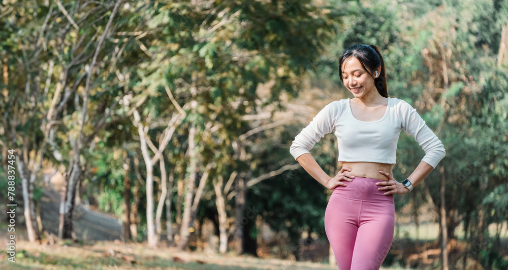 Smiling woman in a white cropped top and pink leggings stands with hands on hips on a paved park pathway, trees in the background. Smiling woman in a white cropped top and pink leggings stands with hands on hips on a paved park pathway, trees in the background.