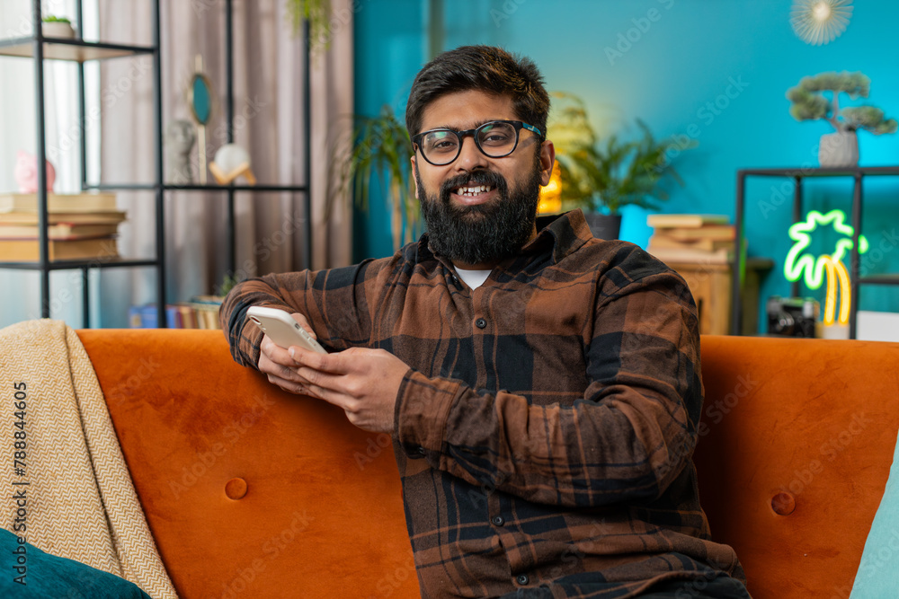 Happy smiling man in casual clothes thinking and text messaging on smartphone. Indian guy sitting on sofa and using cellphone in living room at home. Male is searching information on browser internet. Happy smiling man in casual clothes thinking and text messaging on smartphone. Indian guy sitting on sofa and using cellphone in living room at home. Male is searching information on browser internet.