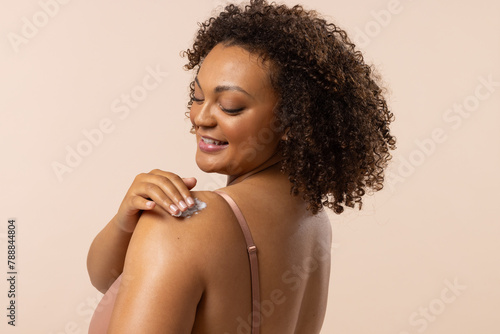 Biracial young female plus size model rubs lotion on shoulder, on beige background, copy space photo