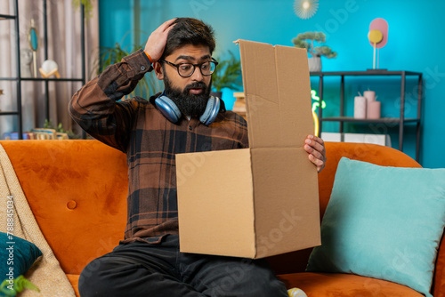 Angry dissatisfied shopper Indian man unpacking parcel feeling upset and confused with wrong mistake delivery from an online store, bad quality broken purchase at home. Guy indoors in room on couch
