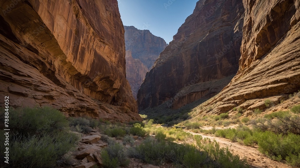 Narrow dirt path winds through canyon, flanked by towering rock walls bathed in warm sunlight. Rugged terrain sparsely dotted with desert vegetation, clear blue sky peeks through canyon's opening.