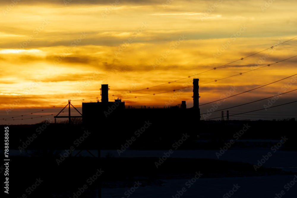 Sunsetting over industrial facility. Silhouette of large refinery with tall exhaust towers.  Power lines reach to the factory. Burning orange sky makes the background