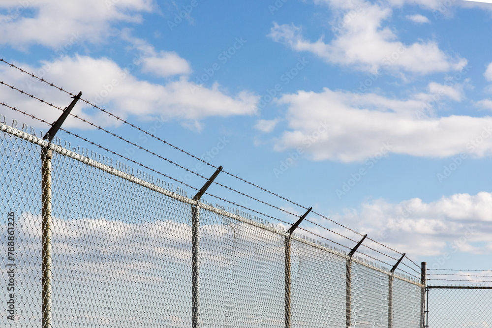Barb wire on top of a chain link fence against a blue cloudy sky