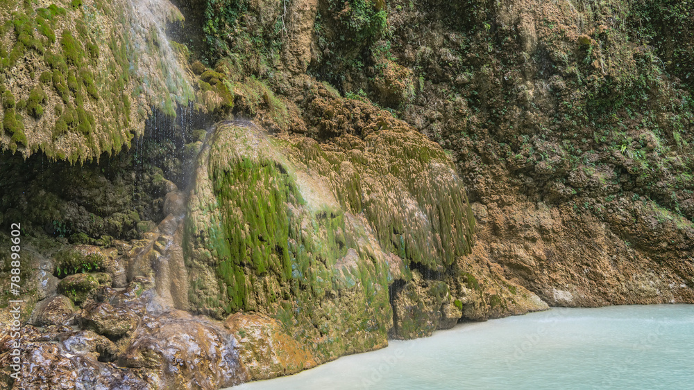 Fototapeta premium A beautiful tropical waterfall. Thin streams of water flow down the mossy stone terraces of the slopes, forming a veil. Turquoise lake at the foot of the cliff. Philippines. Cebu. Tumalog Falls.