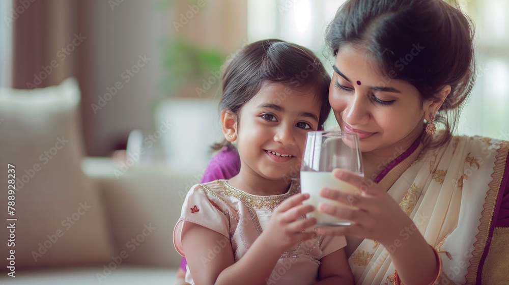 Indian mother in traditional clothes sari, little daughter holding glass of milk, sitting on sofa at home Indian mother in traditional clothes sari, little daughter holding glass of milk, sitting on sofa at home