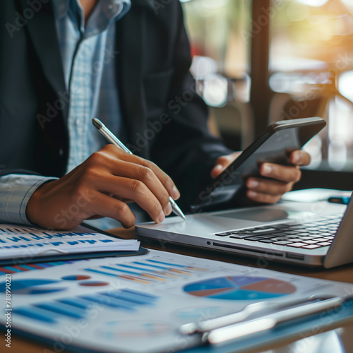 A businessman sits in his office, holds cellphone and a pen, uses a laptop, calculates financial numbers, examines the financial data from the corporate finance chart. Copy space.