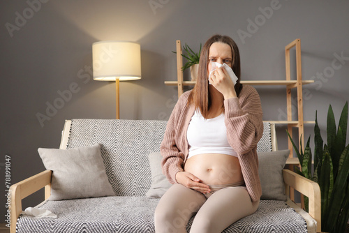 Virus grippe during pregnancy. Unhealthy pregnant woman wearing casual clothing sitting on sofa at home catching cold suffering runny nose sneezing using napkin