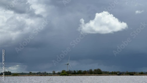 Single windmill on the edge of a lake rotating in storm wind against ominous, dark stom sky, regenerating renewable, sustainable green energy and electricity photo