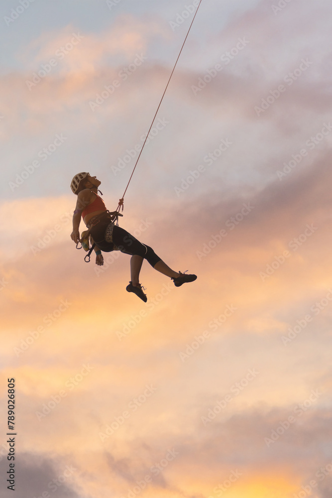 A woman is hanging from a rope, and the sky is orange and pink. Scene is adventurous and exciting A woman is hanging from a rope, and the sky is orange and pink. Scene is adventurous and exciting