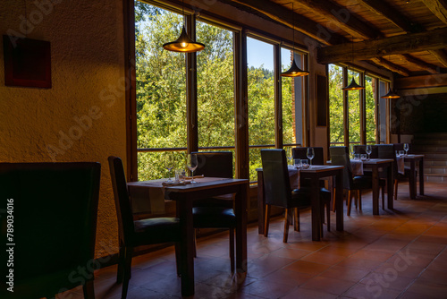 Rustic restaurant room of a mountain hotel, with large windows overlooking the forest and the mountains, luxuriously decorated with wooden ceiling and beams, terrazzo floor and romantic lighting.