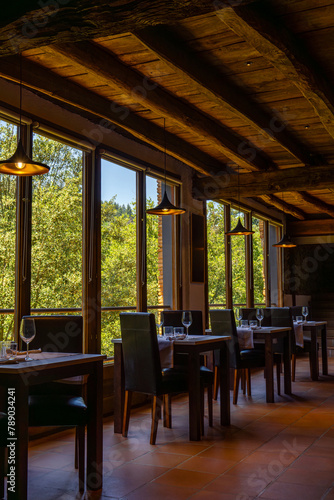 Rustic restaurant room of a mountain hotel  with large windows overlooking the forest and the mountains  luxuriously decorated with wooden ceiling and beams  terrazzo floor and romantic lighting.