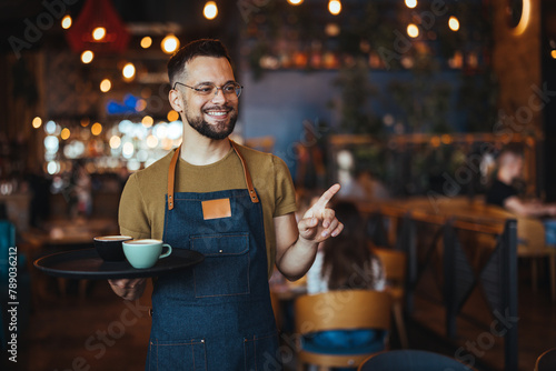 Portrait of smiling young waiter holding tray in cafe. Handsome waiter smiling at camera holding tray at the coffee shop. Profesional Waiter in Restaurant