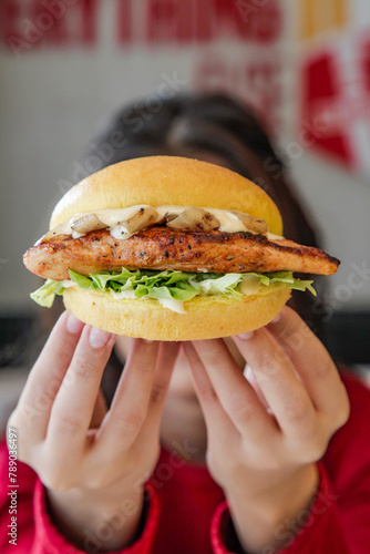 Female holding juicy tender grilled chicken brioche bun burger with hand fingers in front of her face hiding behind.