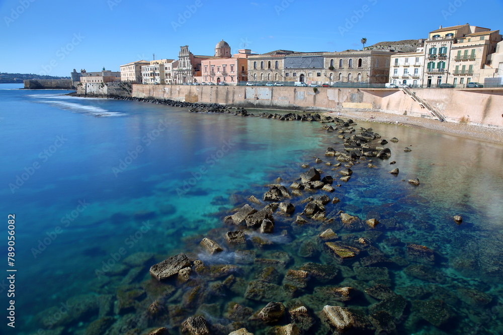 The Eastern rocky coast of Ortigia Island, Syracuse, Sicily, Italy, with clear and colorful water and Castello Maniace (Maniace castle) in the background on the left