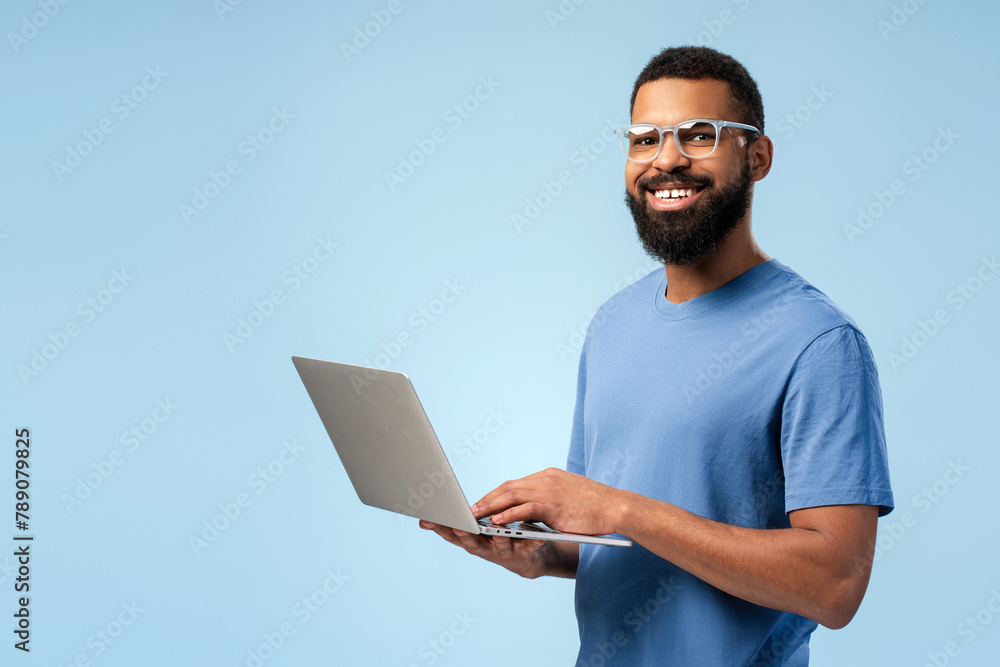Smiling African American man in eyeglasses typing on the laptop while looking at camera Smiling African American man in eyeglasses typing on the laptop while looking at camera