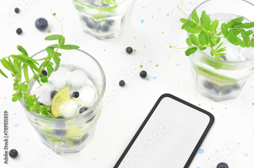 Drinks in glasses and phone with empty space to fill with content. Decorations: leaves and blueberries on light table with little blue paint stains.