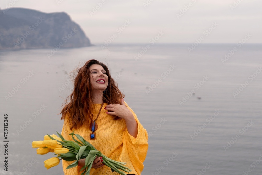 Portrait happy woman woman with long hair against a background of mountains and sea. Holding a bouquet of yellow tulips in her hands, wearing a yellow sweater Portrait happy woman woman with long hair against a background of mountains and sea. Holding a bouquet of yellow tulips in her hands, wearing a yellow sweater