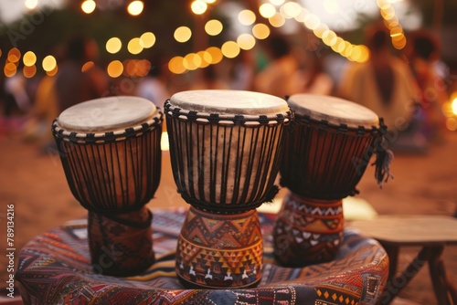 Three traditional African drums placed on an embroidered cloth, with a blurred background of a cultural event and bokeh lights, with people dancing in the distance. The drum heads have intricate photo