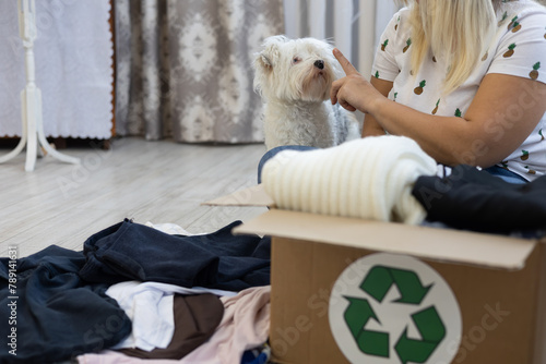 A woman plays with her dog while assembling clothes for recycling. The concept of sorting clothes to be sent for recycling. A box with a recycling sign is on the floor next to the woman.