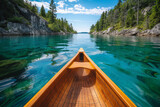The bow of a wooden canoe navigates through the clear waters of a serene lake, flanked by rugged cliffs and lush greenery under a vibrant blue sky.