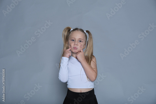 Child making a funny face, her pigtails adding to the silly, adorable gesture, standing against a plain background.