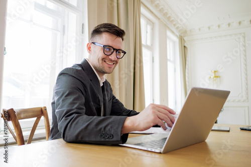 A content manager man with stylish glasses works intently on a laptop at a spacious table in a room graced with classical detailing.