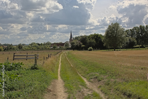  Footpath along fields with trees in the Flemish countryside with church in the distance on a cloudy summer day  photo