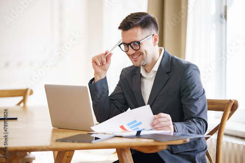 An upbeat businessman analyzes data charts, poised with pen in hand, at a worktable with a laptop, emanating positivity and focus.