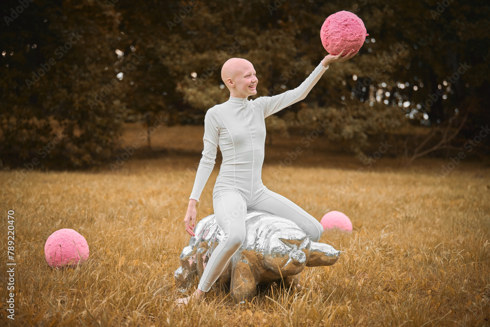 Young hairless girl with alopecia in white cloth sits on tardigrade figure and holds pink ball in hand on fall lawn park, surreal scene with bald teenage girl engages with symbolic elements - obrazy, fototapety, plakaty 