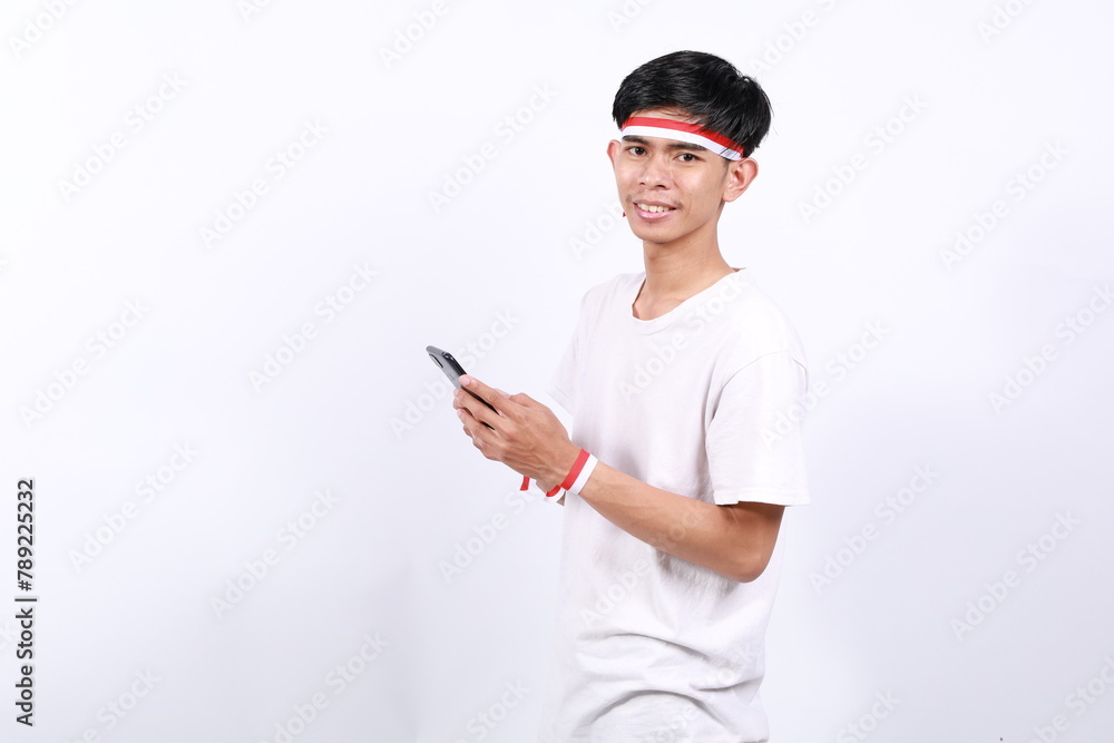 Happy Asian young boy with red white ribbon for celebrating Indonesia independence day standing looking at the camera while holding cell phone Happy Asian young boy with red white ribbon for celebrating Indonesia independence day standing looking at the camera while holding cell phone