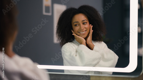 Happy smiling satisfied African American woman looking in mirror feeling joyful pampering in morning bathrobe smile touching face and curly hair girl female skin care haircare cosmetics getting ready