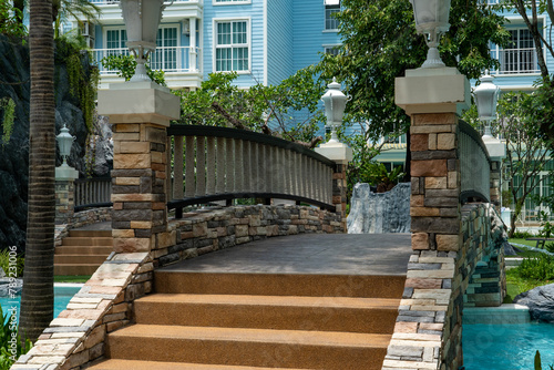 A stone bridge with decorative lanterns and steps spans a blue water pool in a condominium in a tropical resort.	