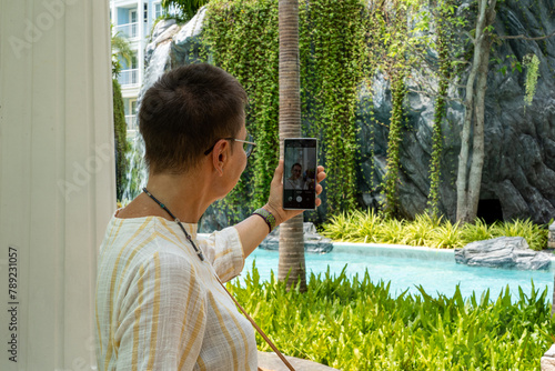 An elderly woman in glasses takes a selfie on her phone in the courtyard of a condominium while standing next to the swimming pool.