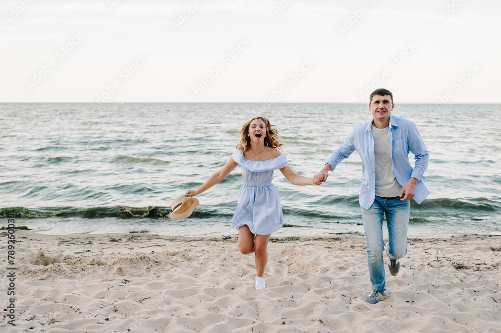 Happy couple in love holding hands and looking at camera on seashore. Man and woman running on sand sea. Female and male run on beach ocean and enjoying summer day on vacation. Spending time together.