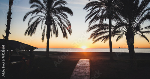 Serene beach sunset Spain with wooden pathway, palm trees, and distant ships. Perfect maritime evening imbued with tranquility and warmth