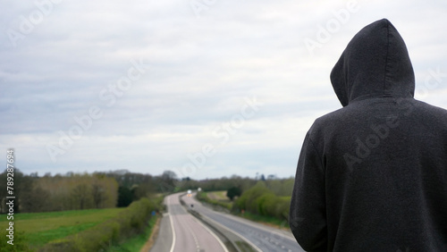 A mysterious hooded figure, back to camera. Looking down on an empty motorway in the countryside. With copy space