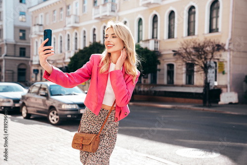 Young beautiful smiling blond hipster woman in trendy summer jacket clothes. Sexy carefree female posing in the street at sunny day. Positive model outdoors at sunset. Cheerful and happy, takes selfie