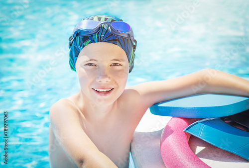 Active, happy child (boy) in cap, sport goggles ready to learns professional swimming with pool board, swim noodles and fins for swimming. Kid enjoying water in swimming pool. 