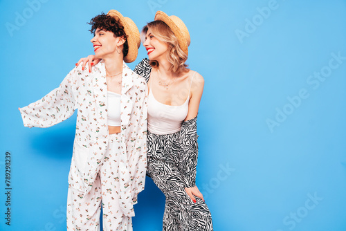 Two young beautiful smiling brunette hipster female in trendy summer costume clothes. Sexy women posing near blue wall in studio. Positive models having fun. Cheerful and happy. In hat, red lips