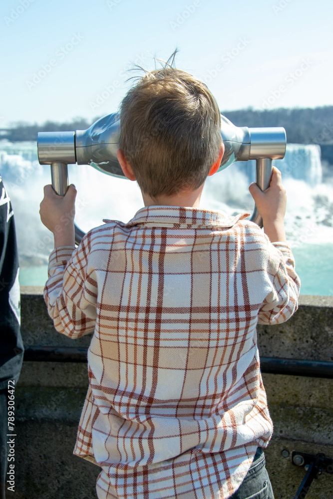 Boy looking through the viewing binoculars on the balcony across the American and Bridal Veil Falls crashing over rocky cliff. Tourist observing wild whitewater waterfalls on Niagara River. High